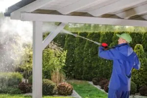 A person in blue rain gear uses a pressure washer to clean the roof of a wooden structure, surrounded by greenery and bushes, showcasing the benefits of pressure washing for your home's exterior.