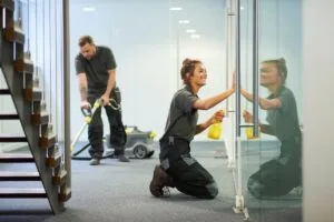 Two people in work uniforms demonstrate how to maintain a germ-free office: one vacuums the carpet while the other meticulously wipes a glass wall with a spray bottle.