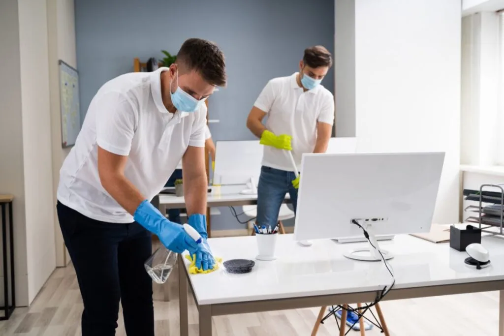 Two people wearing masks and gloves demonstrate how to maintain a germ-free office by cleaning white desks with spray bottles and cloths.