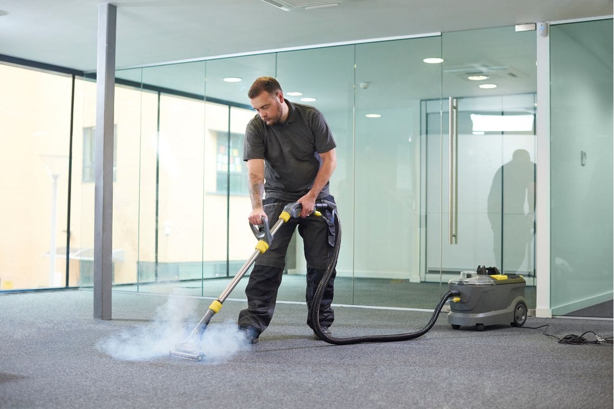 A man demonstrates how to maintain a germ-free office by using a steam cleaner on an office carpet, surrounded by glass walls that reflect the pristine environment.