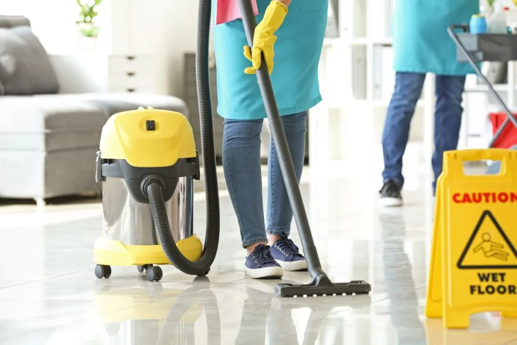 Wearing blue jeans and gloves, a person diligently vacuums the shiny floor, ensuring it's not just spotless but also germ-free. The yellow caution wet floor sign stands nearby as a reminder of how to maintain a hygienic office environment.
