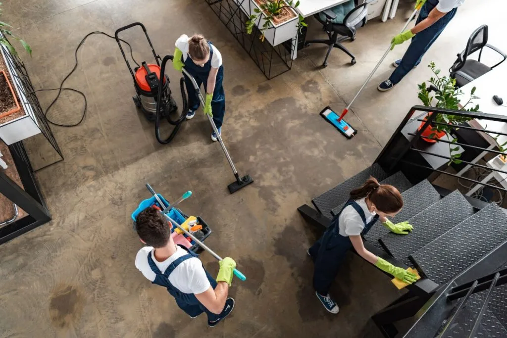 Four people wearing uniforms and gloves diligently clean an industrial-style office space, showcasing how to maintain a germ-free office. They use vacuums, mops, and various cleaning tools on the floor and stairs to ensure optimal hygiene.