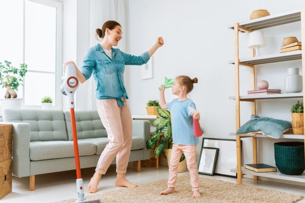A woman and a child dance joyfully with cleaning tools in hand, embracing tips for a cleaner home this spring. The scene is set in a lively living room with a vacuum and spray bottle, pulsating to the rhythm of tidy energy.