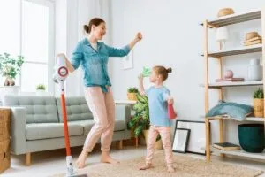 A woman and a child dance joyfully with cleaning tools in hand, embracing tips for a cleaner home this spring. The scene is set in a lively living room with a vacuum and spray bottle, pulsating to the rhythm of tidy energy.
