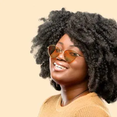 Woman with curly hair wearing sunglasses and a beige sweater smiles against a light background.