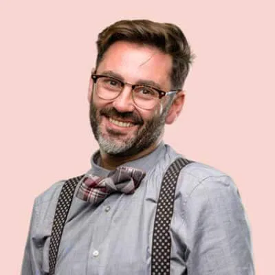 A man with glasses and a beard is smiling. He is wearing a light blue shirt, a plaid bow tie, and suspenders. The background is a solid light pink.