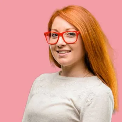 A person with long red hair and red glasses is smiling, wearing a light-colored sweater against a pink background.