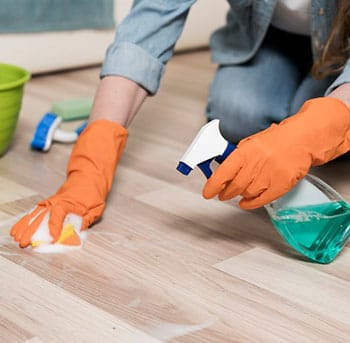 Person wearing orange gloves cleans a wooden floor with a sponge and spray bottle.