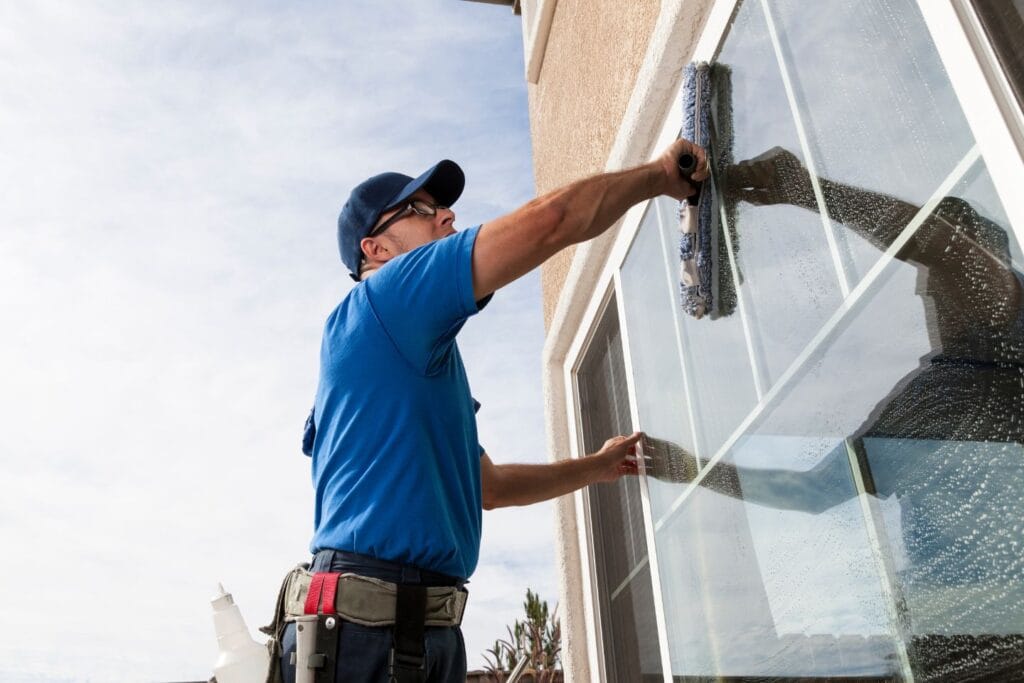 A person in a blue uniform and cap is diligently cleaning a large home window with a squeegee on the building's exterior.