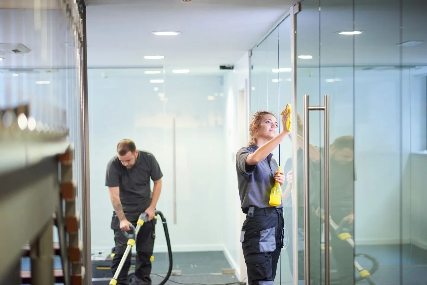 Two cleaners work in an office that feels almost like home; one vacuums the floor while the other cleans the glass door.