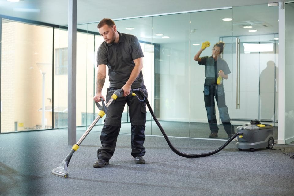 A person is diligently vacuuming a carpet in an office with glass walls, showcasing the efficiency of commercial cleaning, while another person carefully cleans the glass in the background.