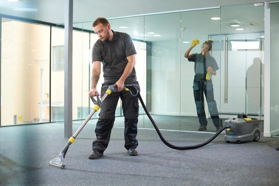 A person is diligently vacuuming a carpet in an office with glass walls, showcasing the efficiency of commercial cleaning, while another person carefully cleans the glass in the background.
