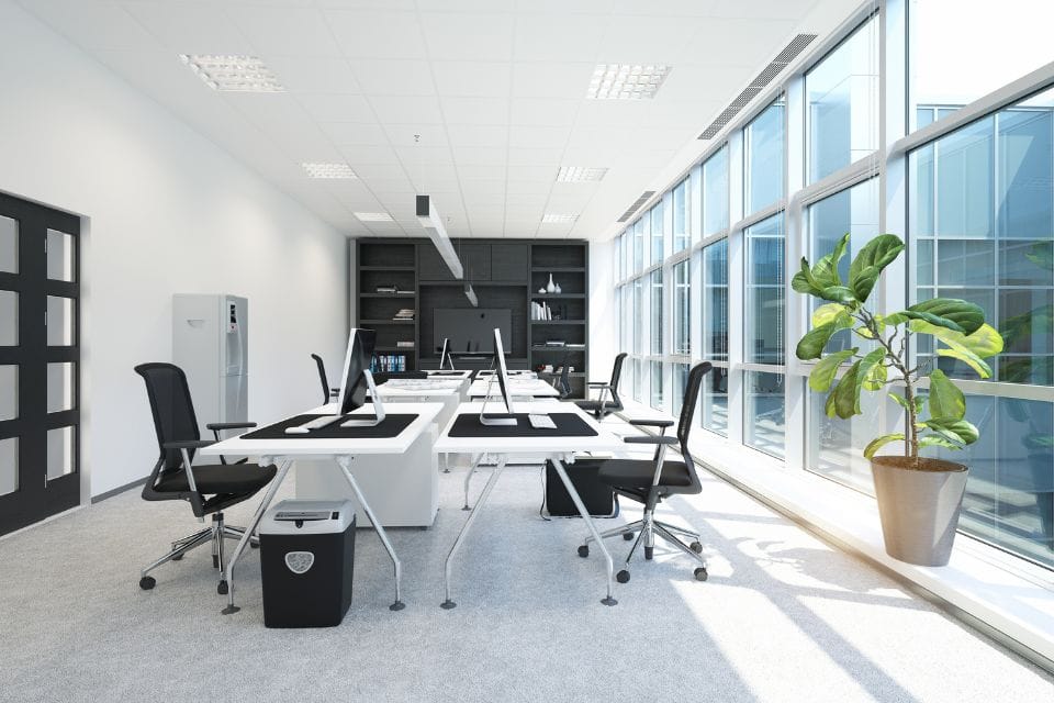 Modern office space with two desks, swivel chairs, and computers under a large window. A potted plant adds a touch of nature while the shelves in the background remain dust-free, thanks to regular commercial cleaning.