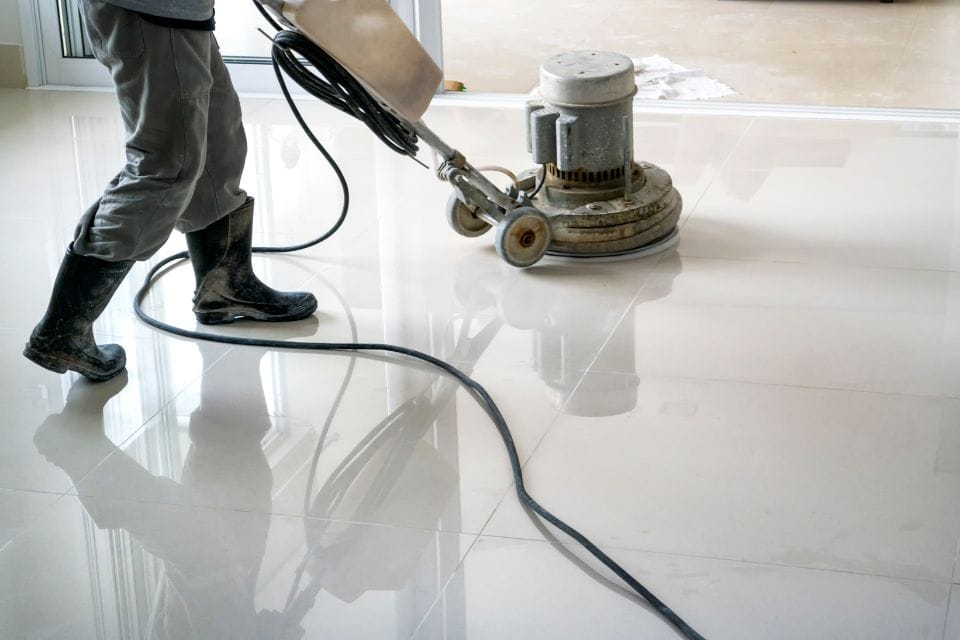 A person is engaged in commercial cleaning, expertly polishing a large, white tile floor with a floor buffer. They sport rubber boots and confidently grip the machine's handle as it glides over the surface.
