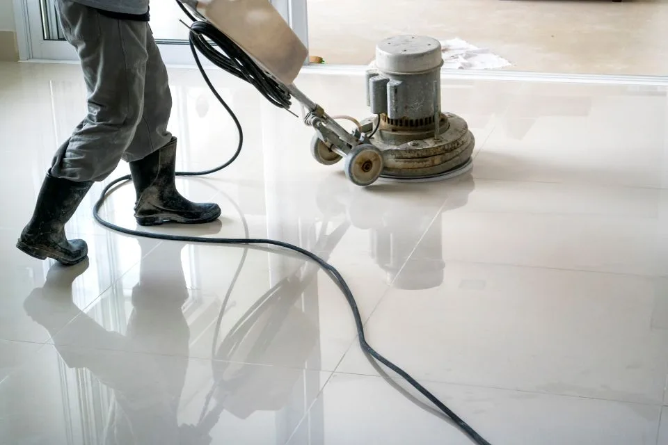 A person is engaged in commercial cleaning, expertly polishing a large, white tile floor with a floor buffer. They sport rubber boots and confidently grip the machine's handle as it glides over the surface.