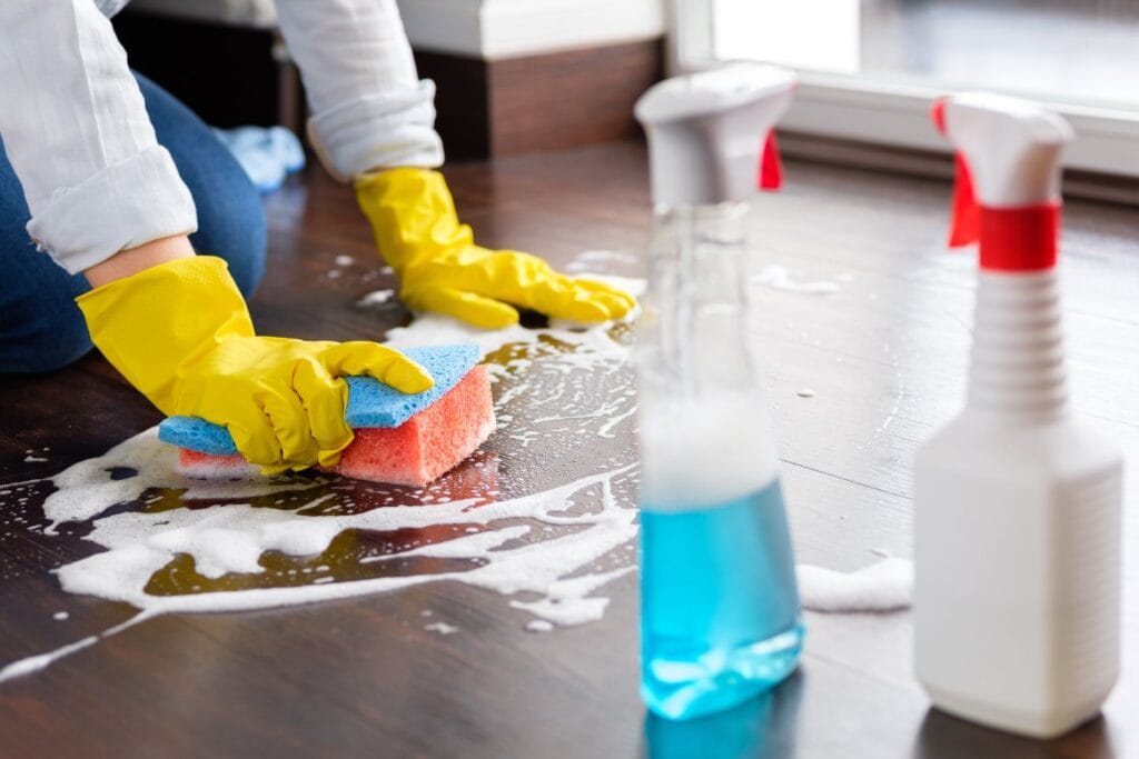 Person wearing yellow gloves scrubbing a soapy wooden floor with a sponge, engaged in deep cleaning. Two cleaning spray bottles are visible in the foreground.