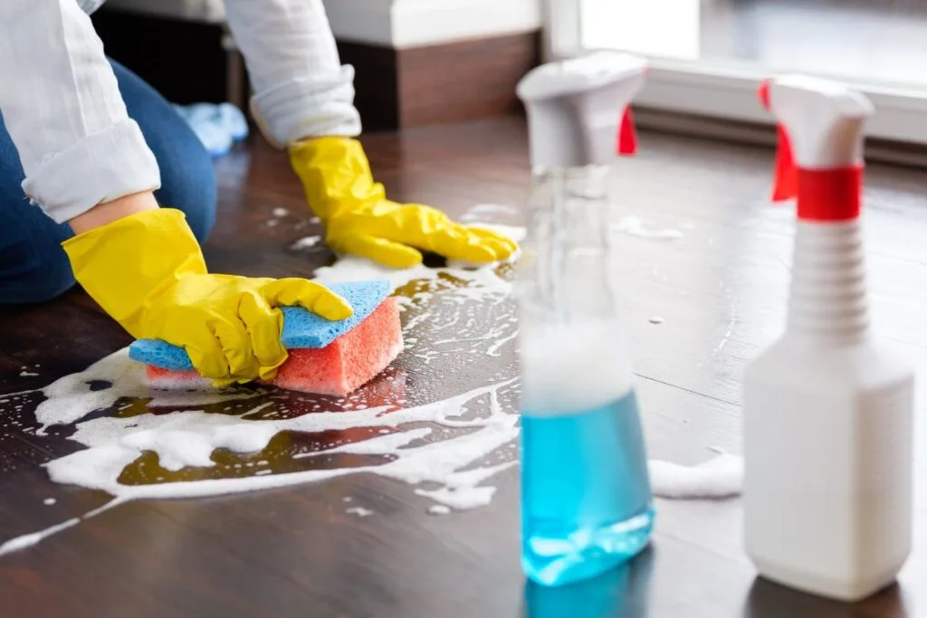 Person wearing yellow gloves scrubbing a soapy wooden floor with a sponge, engaged in deep cleaning. Two cleaning spray bottles are visible in the foreground.