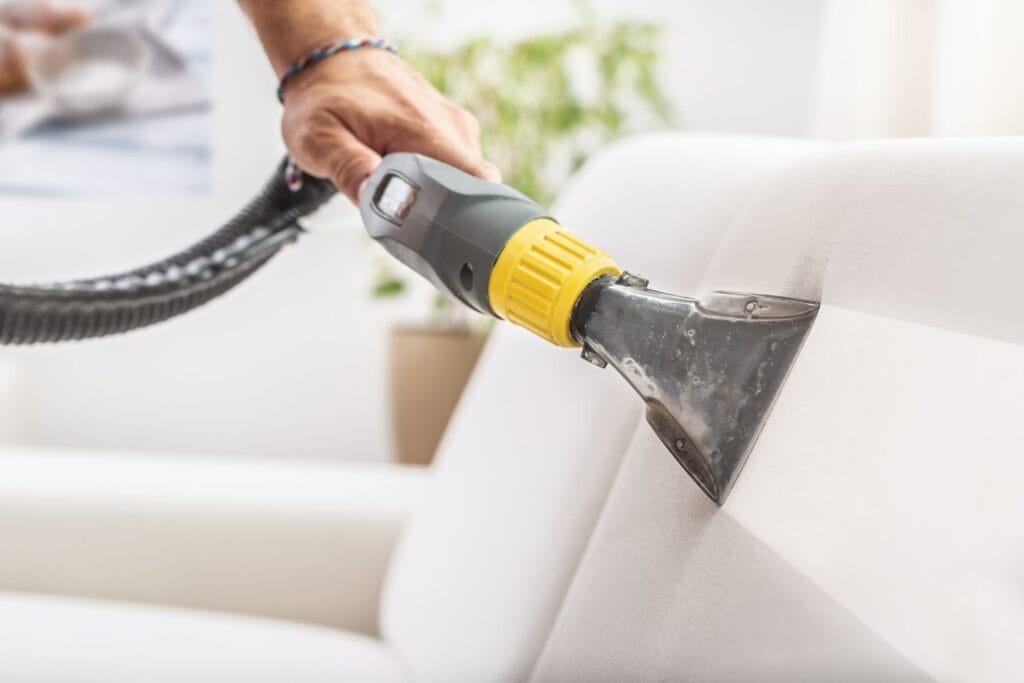 A person is deeply cleaning a light-colored sofa using an upholstery cleaner, ensuring every inch sparkles and shines.