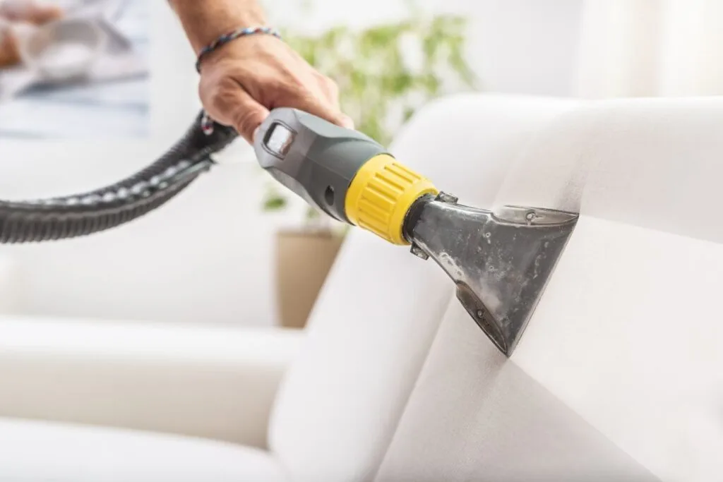 A person is deeply cleaning a light-colored sofa using an upholstery cleaner, ensuring every inch sparkles and shines.
