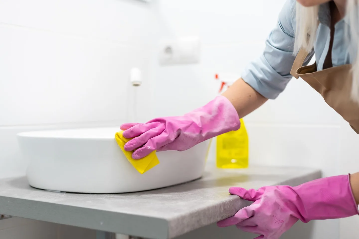 A person in pink gloves meticulously performs a deep cleaning of the bathroom sink using a yellow cloth.