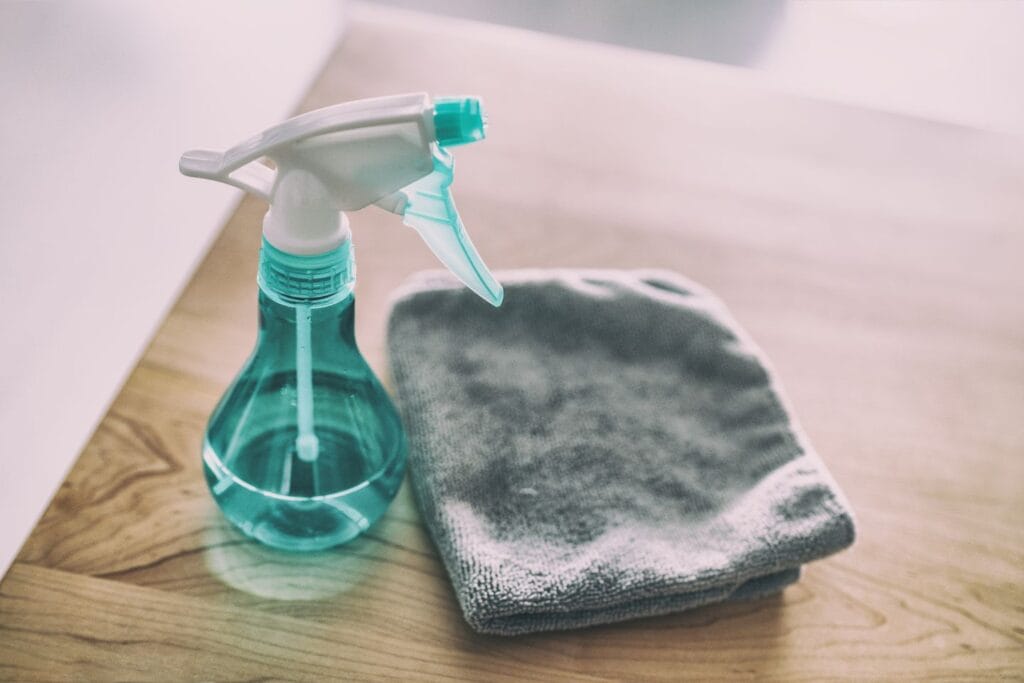 A spray bottle filled with a blue liquid, ideal for disinfecting, is placed next to a folded gray cloth on a wooden surface.