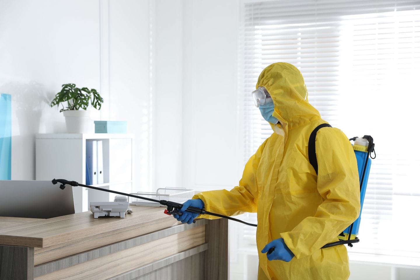 A person in a yellow protective suit is meticulously sanitizing an office desk with a sprayer, highlighting the importance of disinfecting services in maintaining a clean and safe work environment.