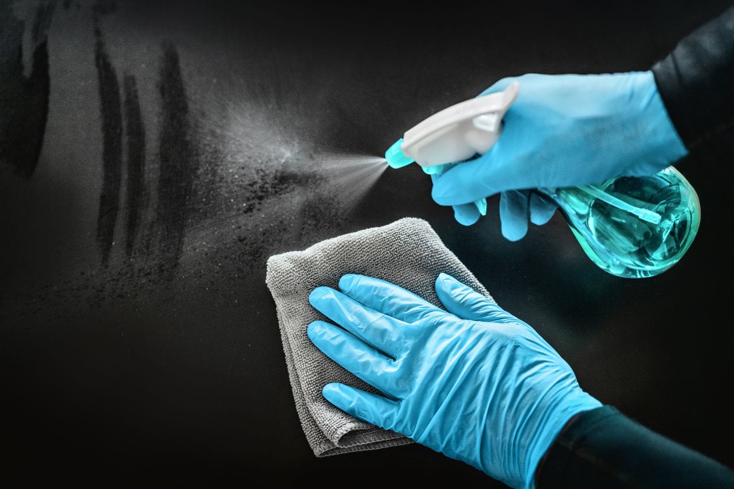 Hands wearing blue gloves spray cleaning solution and wipe a surface with a gray cloth, showcasing professional disinfecting services.
