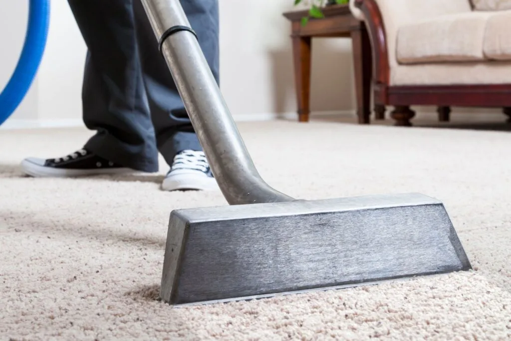 A person is tidying up their home, vacuuming a beige carpet with a metal vacuum head, dressed in black pants and sneakers. In the background, you can see the cozy setup of a brown sofa and a wooden table.