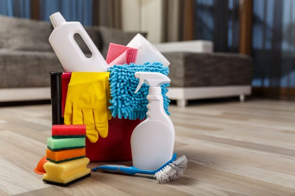 A red cleaning caddy filled with supplies—sponges, a spray bottle, vibrant yellow gloves, a brush, and a mop—sits on the wooden floor of a cozy home, surrounded by stylish furniture.