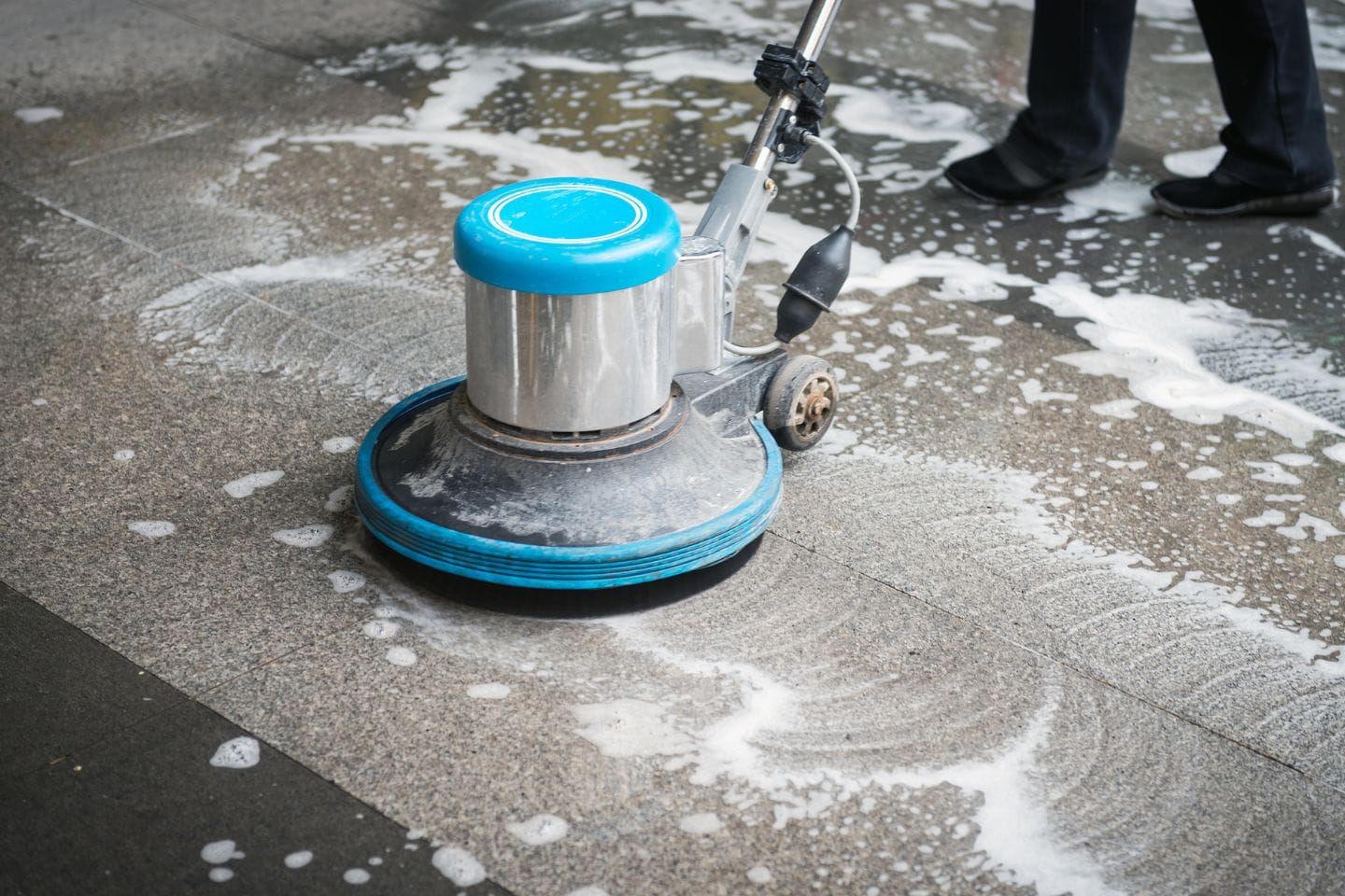A floor cleaning machine scrubs a textured surface in a cozy home, spreading soap suds. A person&rsquo;s legs are partially visible as they deftly operate the machine.
