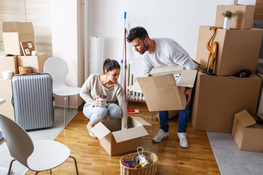 Two people efficiently packing boxes in a room with a wooden floor, surrounded by various items like a suitcase, plants, and a basket, preparing for move-out cleaning to leave the space spotless.