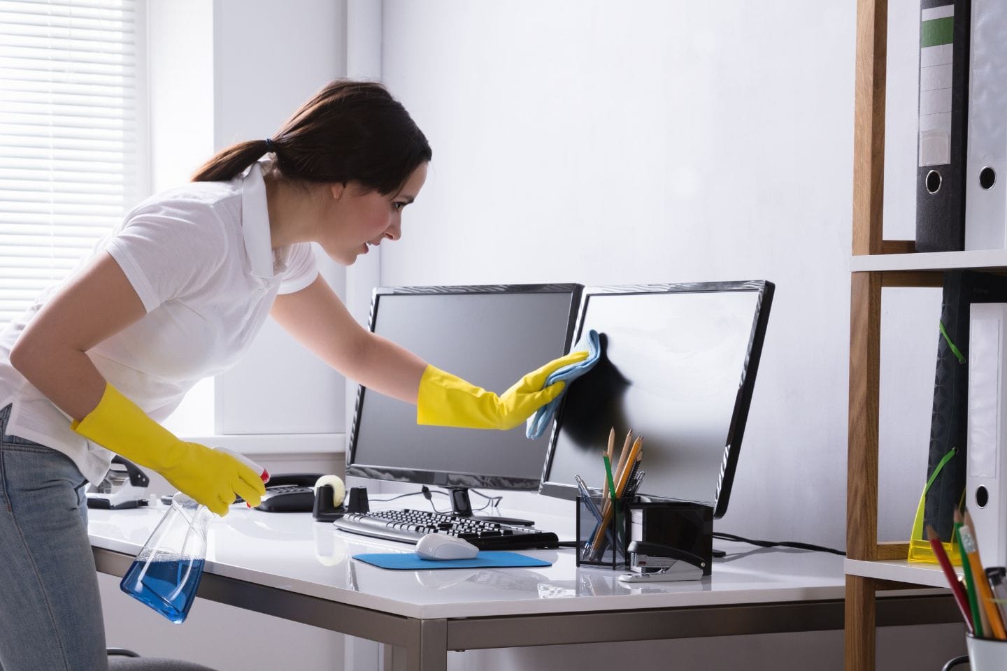 Wearing yellow gloves, the individual engages in meticulous office cleaning, using a cloth and cleaning spray to refresh a computer monitor on a desk filled with office supplies and two monitors.