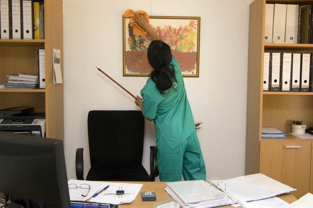 A person in green cleaning attire tidies up a framed painting, efficiently handling office cleaning tasks amid shelves with binders and a desk with supplies. A black chair sits prominently in the foreground, completing the well-organized scene.