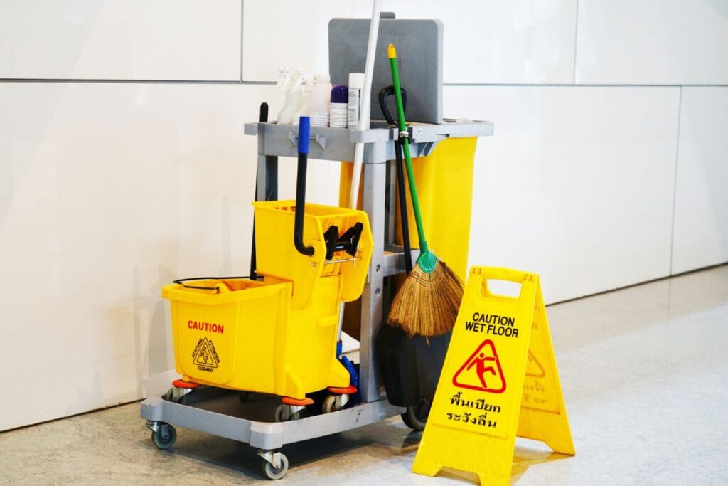 A janitorial cart stocked with office cleaning supplies, including a yellow mop bucket, caution sign, broom, and trash bin, stands ready in the hallway.
