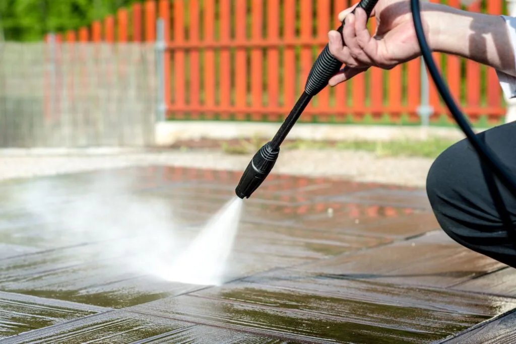 A person expertly employing pressure washing techniques rejuvenates wooden deck tiles, with a striking red fence serving as the backdrop.