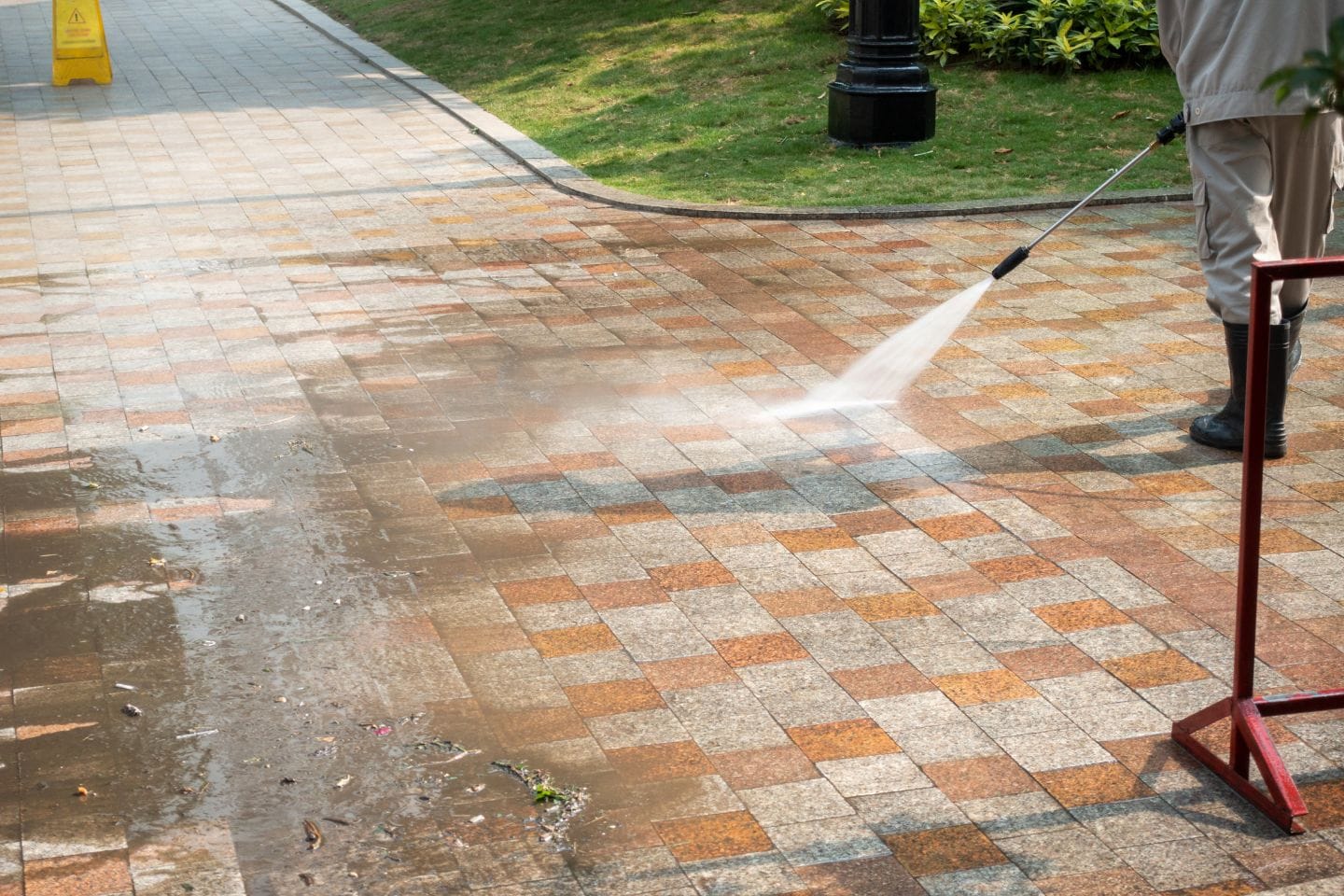 A person expertly pressure washes a brick pathway, with water spraying across the surface, leaving it gleaming. Nearby grass and a warning sign are visible.
