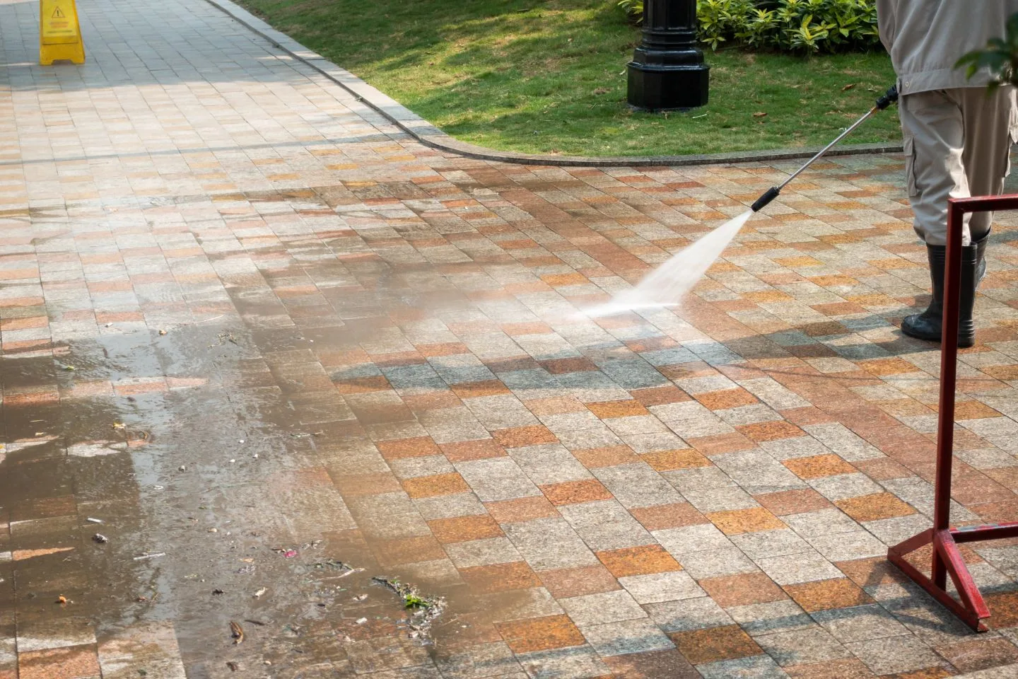 A person expertly pressure washes a brick pathway, with water spraying across the surface, leaving it gleaming. Nearby grass and a warning sign are visible.