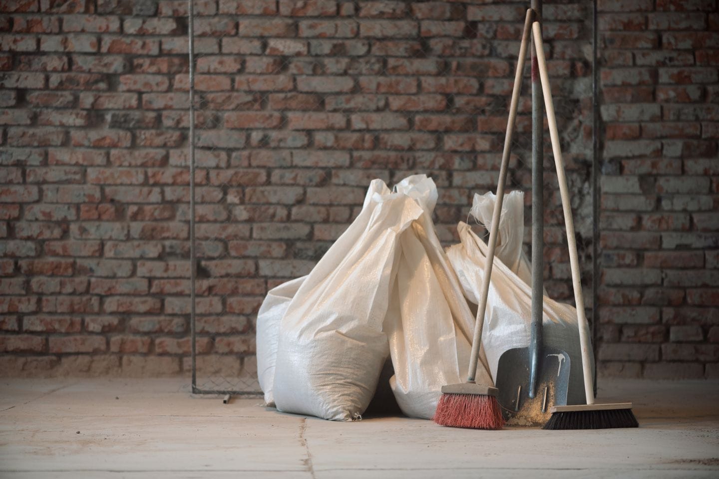 Three large white bags filled with debris stand beside a construction cleanup set featuring a broom, dustpan, and mop against a brick wall backdrop.