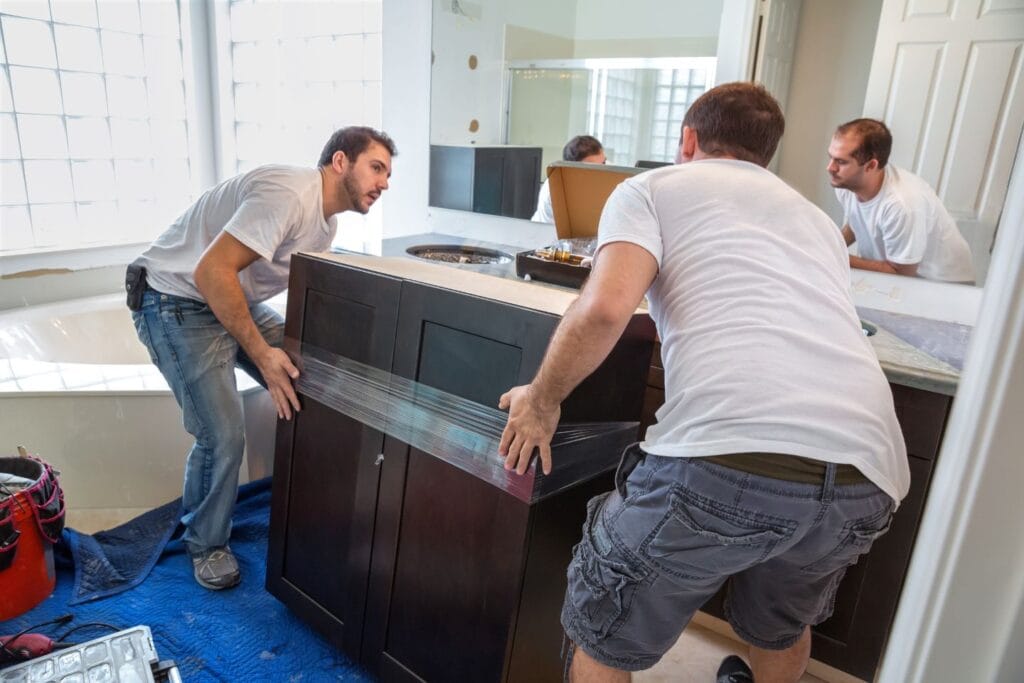 Two men lift and position a wrapped cabinet in a bathroom, preparing for installation. Amidst the post-construction cleanup, both wear casual clothing, and tools are visible nearby.