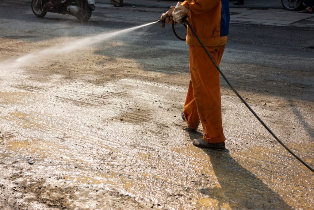 A person in orange clothing is expertly power-washing a concrete surface outdoors, likely as part of post-construction cleaning duties.