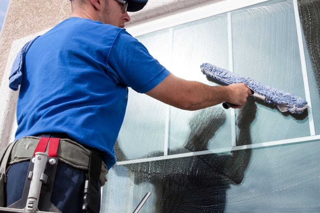 A person in a blue shirt and cap is expertly window cleaning, wielding a squeegee while carrying essential glass cleaning tools in a handy waist belt.