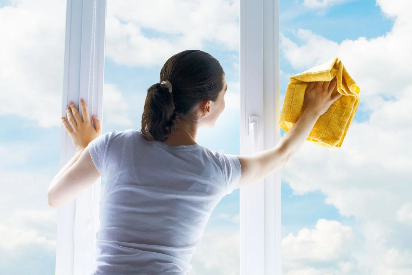 A person in a white shirt diligently engages in window cleaning, using a yellow cloth to polish the glass. Through the gleaming surface, a blue sky adorned with fluffy clouds is visible.