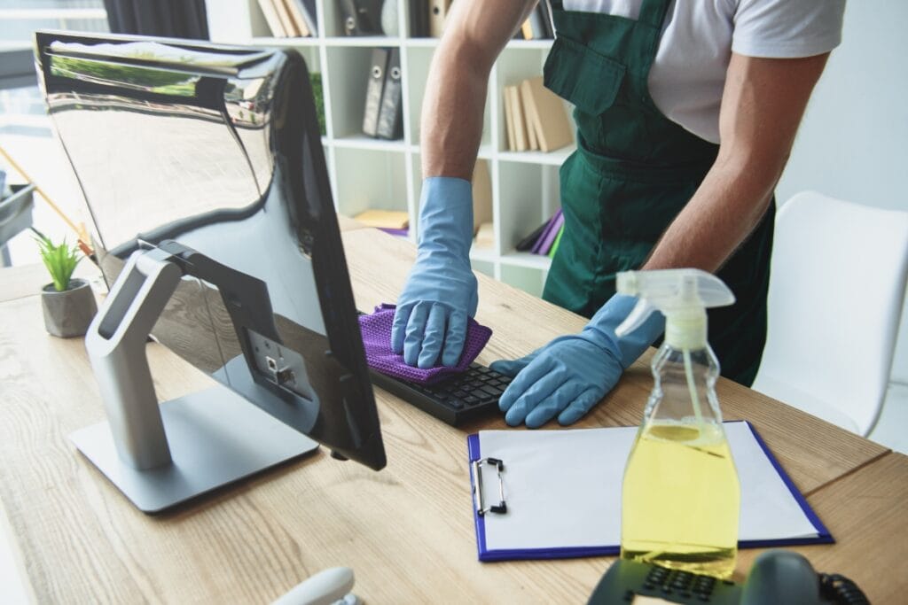 A professional from a commercial cleaning company meticulously cleans a computer keyboard with a purple cloth, sporting blue gloves. Nearby, a spray bottle and clipboard testify to the diligent routine on the desk.