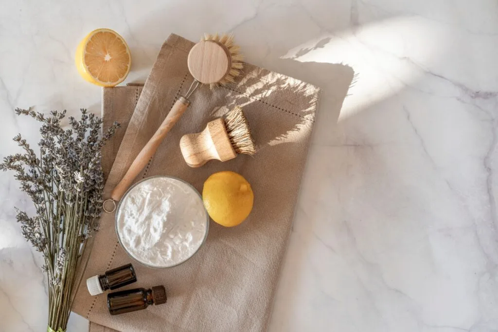 Eco-friendly cleaning products displayed in a natural setup with a bowl of baking soda, lemon, essential oil bottles, dried lavender, and wooden brushes on a linen cloth against a marble background.