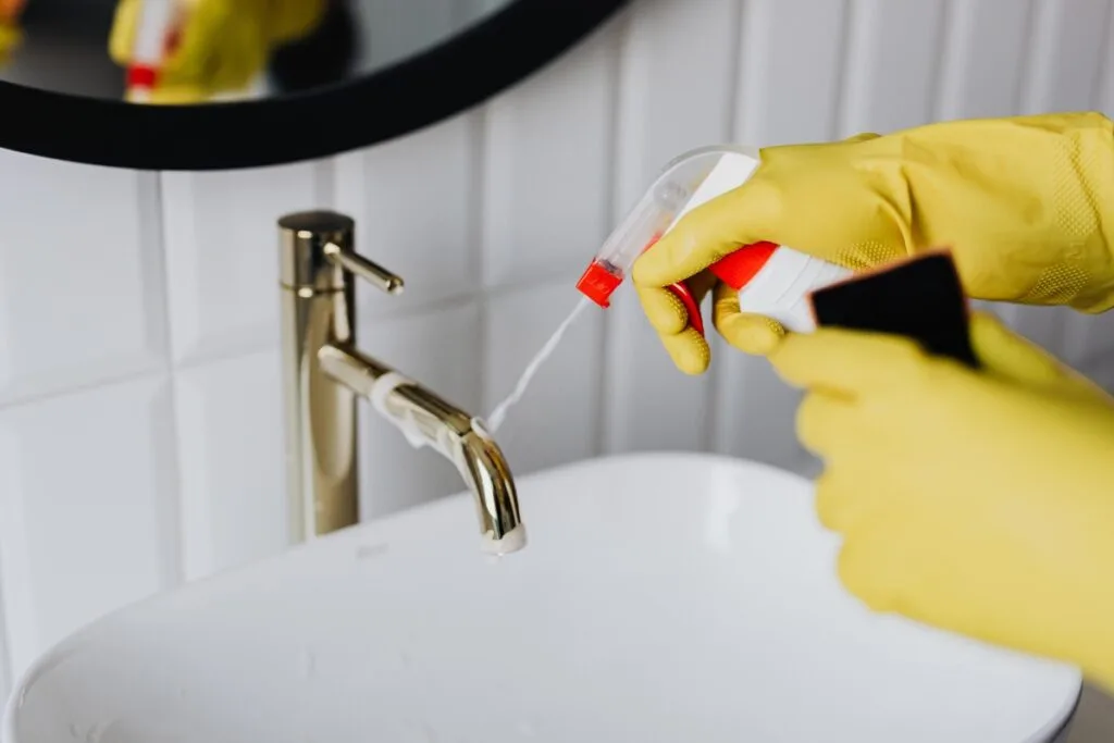 A person wearing yellow gloves sprays an eco-friendly cleaning product onto a bathroom faucet.