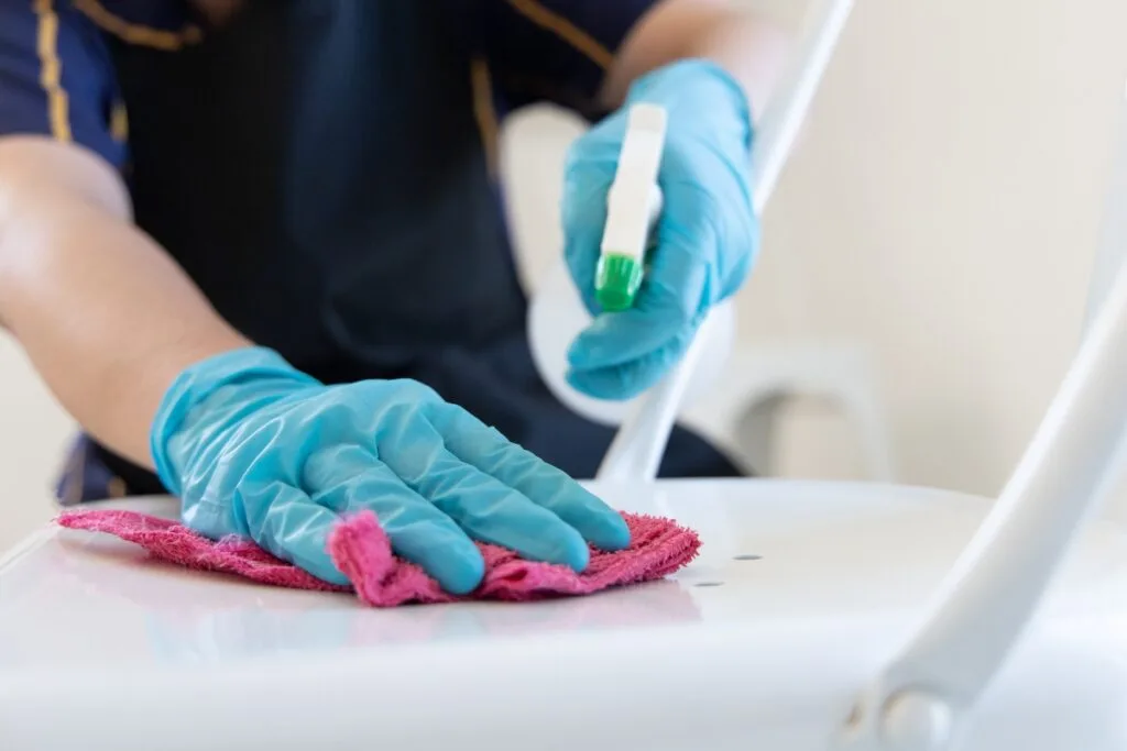 Person wearing blue gloves cleans a white chair with a pink cloth and an eco-friendly spray bottle.
