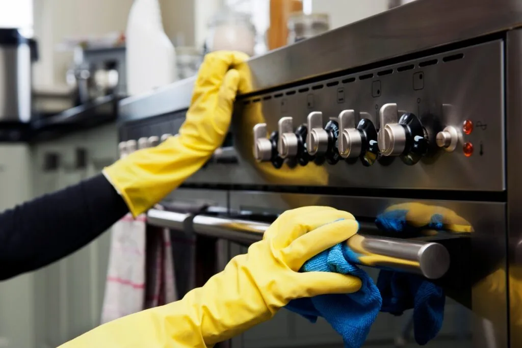 A person wearing yellow gloves uses a blue cloth and eco-friendly cleaning products to shine the stainless steel oven in their kitchen.