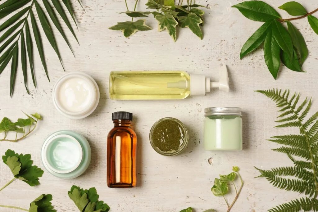 Flat lay of eco-friendly skincare products and glass containers with cream, oil, and gel, surrounded by various green leaves on a light wooden surface.