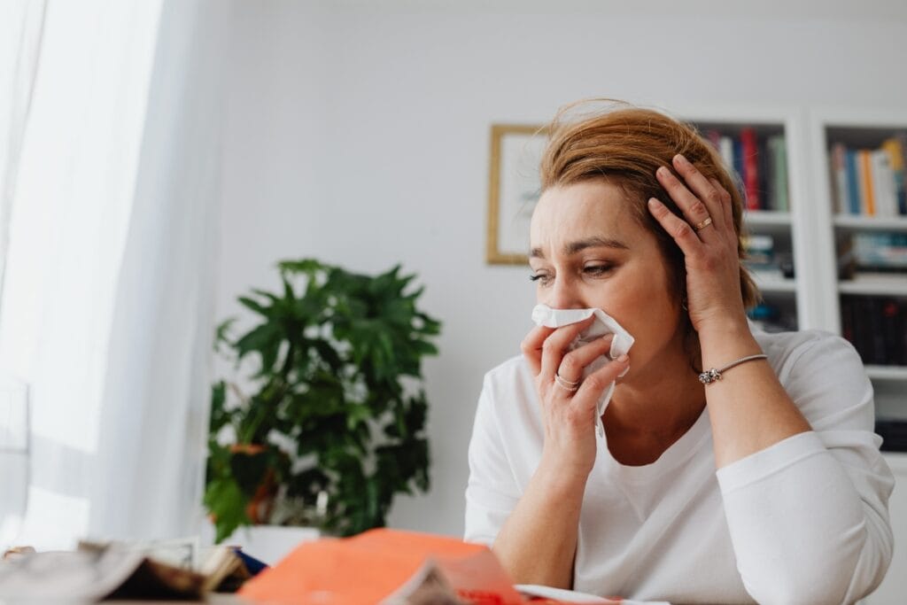 A woman in a white shirt sits at a table, holding a tissue to her nose and appearing unwell&mdash;perhaps affected by allergies&mdash;amid cluttered papers and a plant, highlighting the importance of cleaning for allergy sufferers.