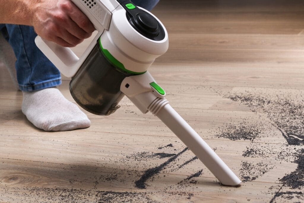 A person uses a cordless handheld vacuum cleaner to clean scattered dirt from a wooden floor, making cleaning for allergy sufferers easier and more effective.
